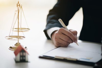 A man is writing notes on paper attached to a clipboard with a balancing scale and a miniature house next to the clipboard, McGrath Law Firm, Real Estate Lawyers, Mount Pleasant, S.C.